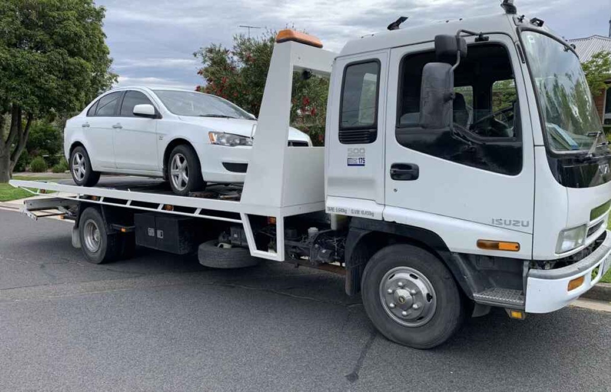 Tow truck operator assisting with battery replacement for a white sedan in West Islip, NY
